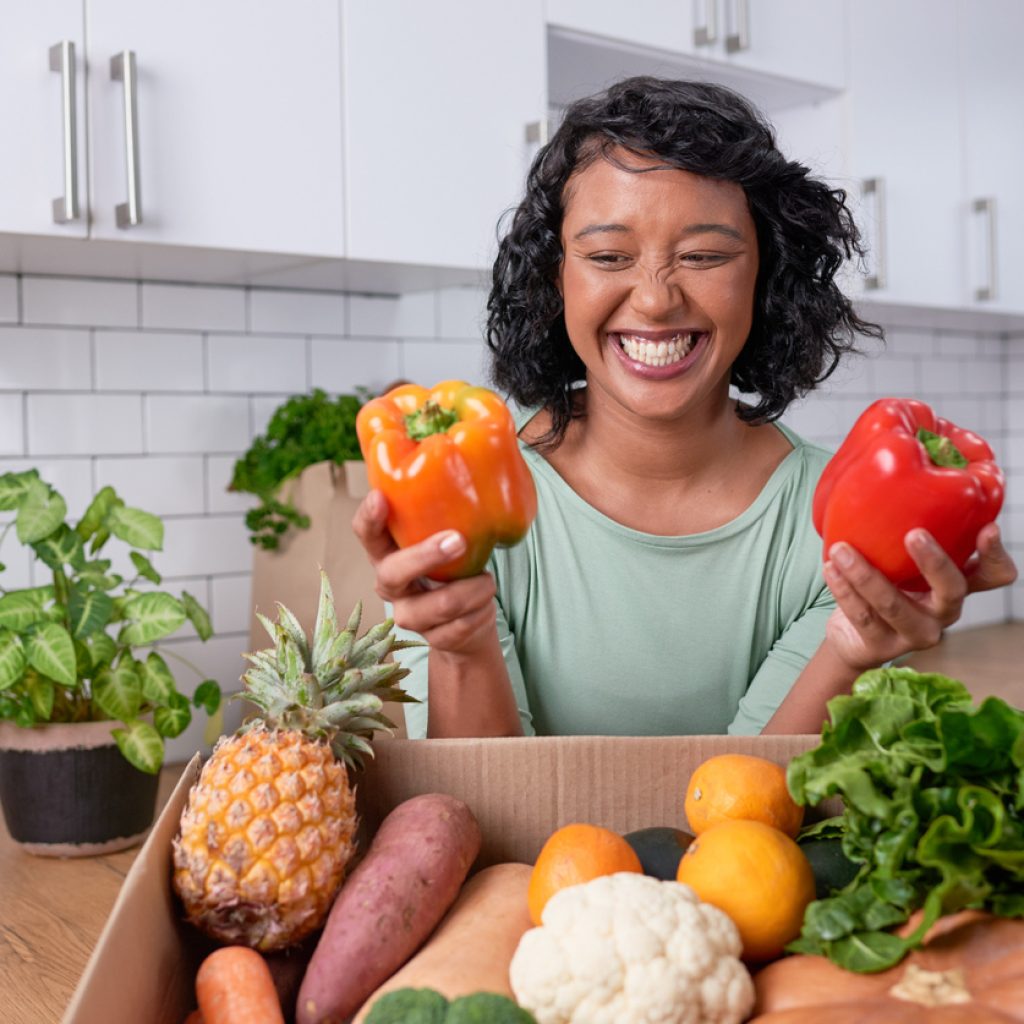 A young multi-ethnic woman holds two sweet peppers out of fresh veg delivery box